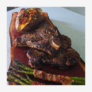 Loaded baked potatoes, two t-bone steaks, and bacon wrapped asparagus plated on a wooded slab