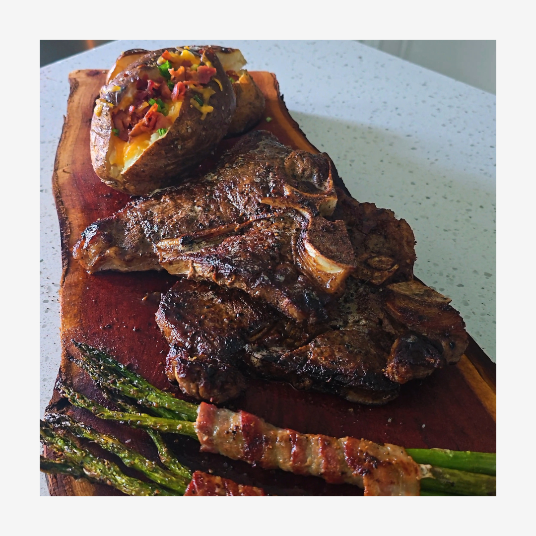 Loaded baked potatoes, two t-bone steaks, and bacon wrapped asparagus plated on a wooded slab